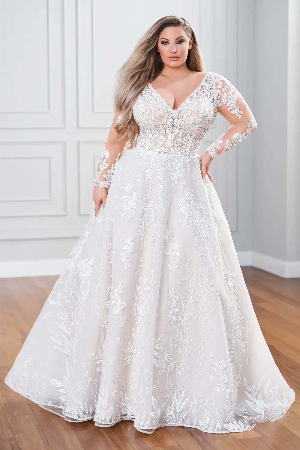 A woman wearing a long-sleeved, elegant white gown with floral embroidery, standing in a bright indoor space.