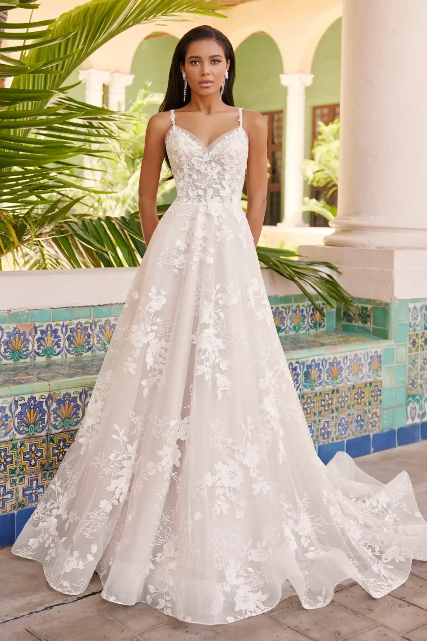 A woman wearing a white, floral lace wedding dress stands in an outdoor setting with colorful tiled walls and plants in the background.