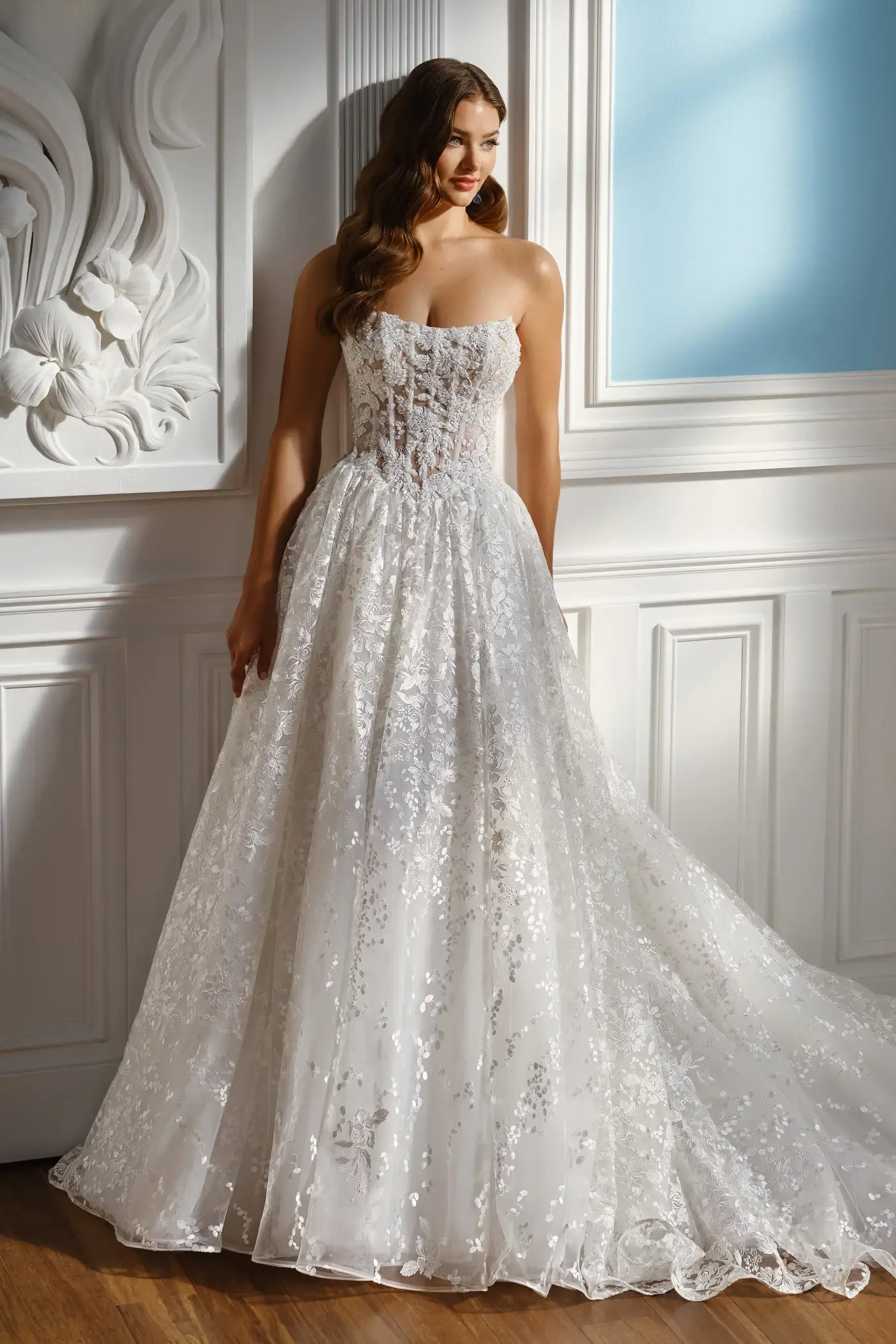 A woman in an elegant, strapless lace wedding gown stands in a bright room. The dress features intricate floral patterns and a fitted bodice, exuding a romantic ambiance.