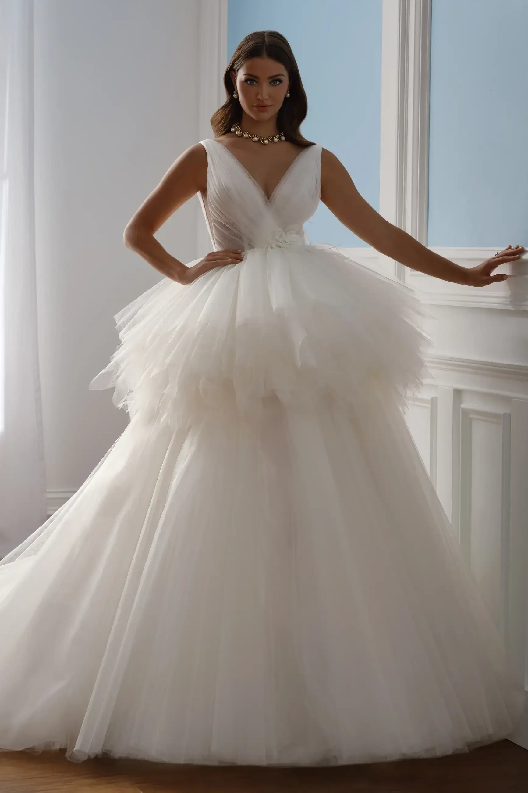 A woman in an elegant white tulle wedding gown stands confidently in a sunlit room. The dress features a deep V-neck and layered, voluminous skirt.