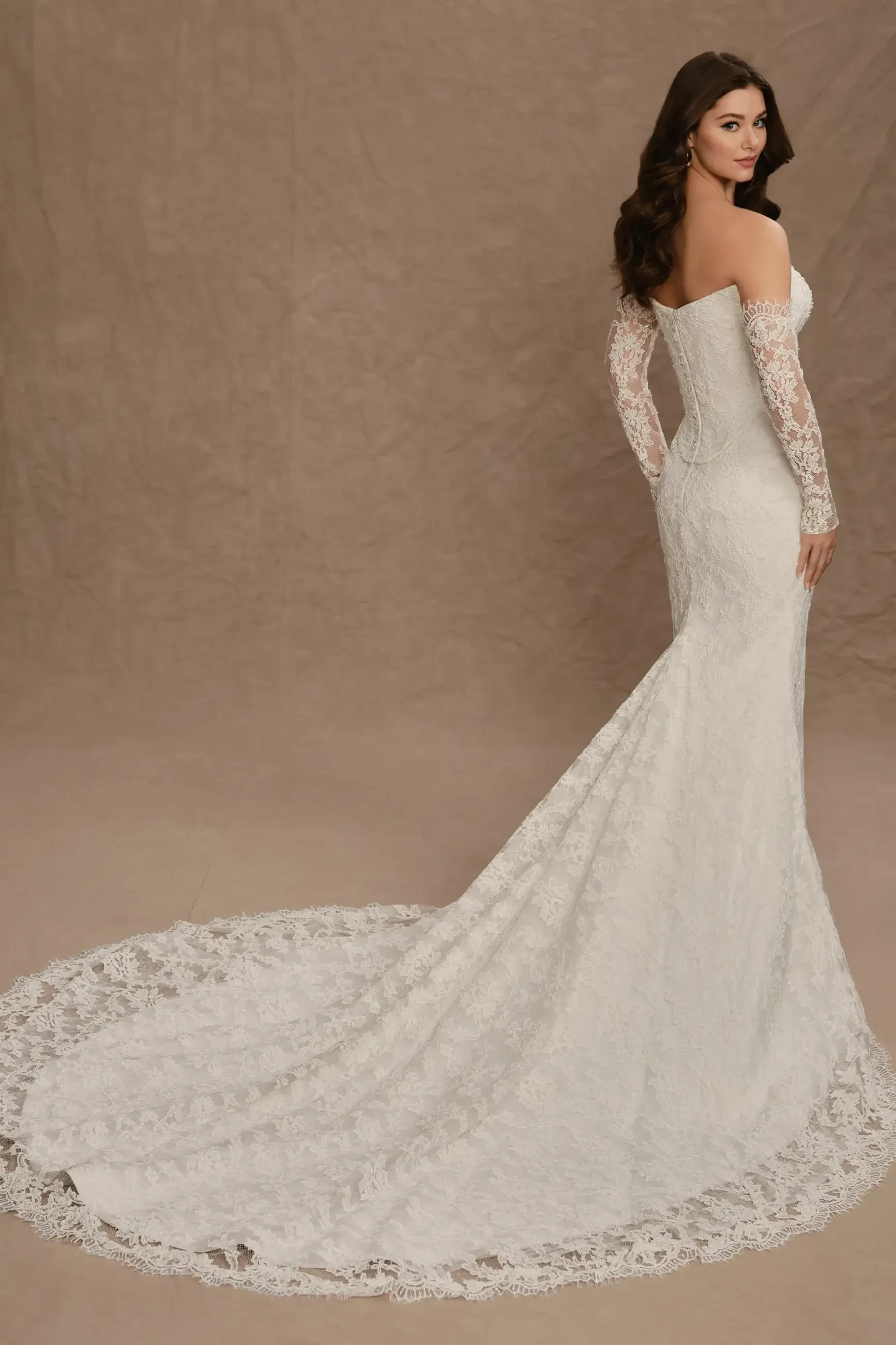 A woman is wearing a beautiful off-the-shoulder wedding dress with lace long sleeves and a flowing train, standing with her back turned against a neutral background.