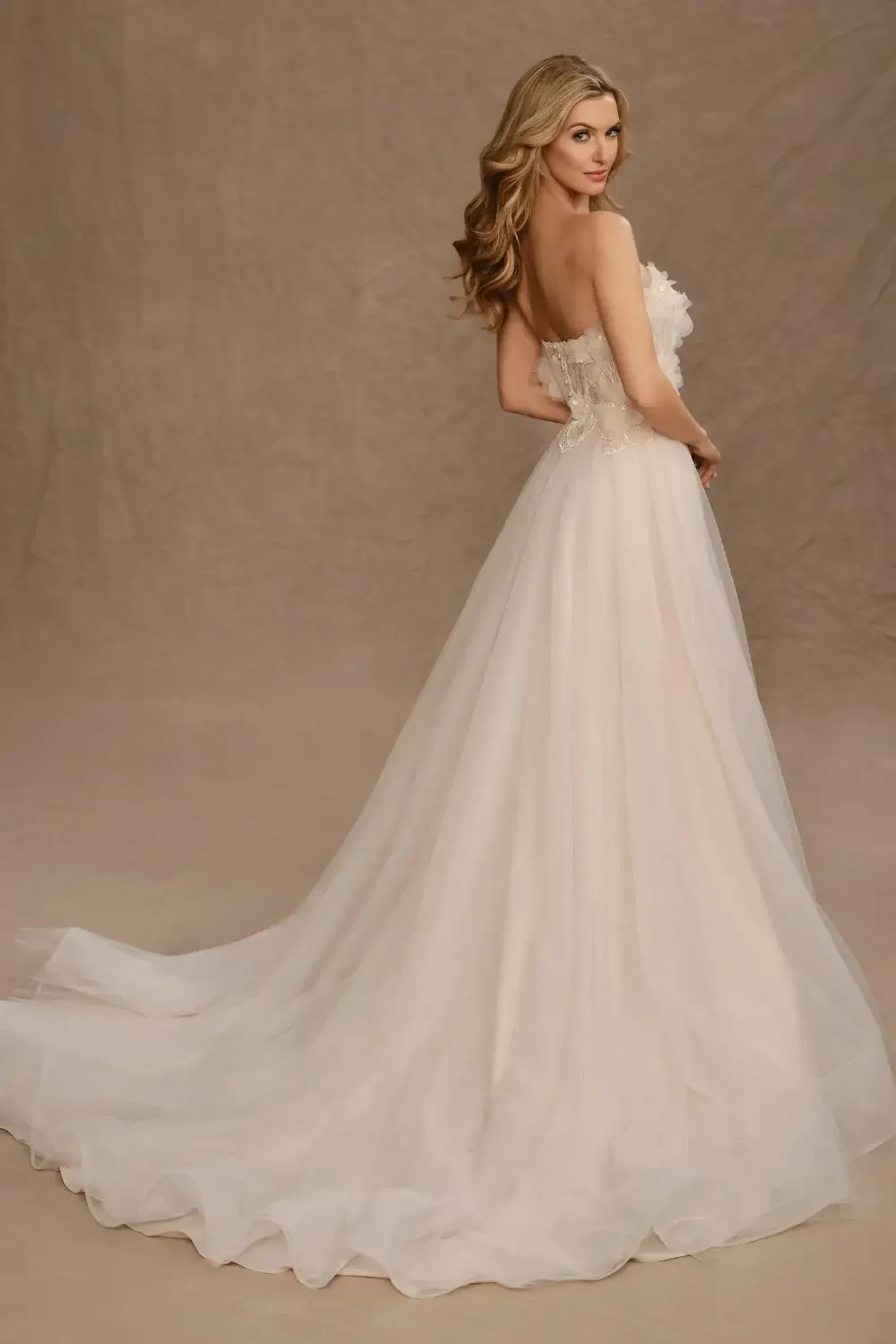 A model in a strapless, flowing gown stands elegantly in a lavish interior with ornate decor and golden lighting.