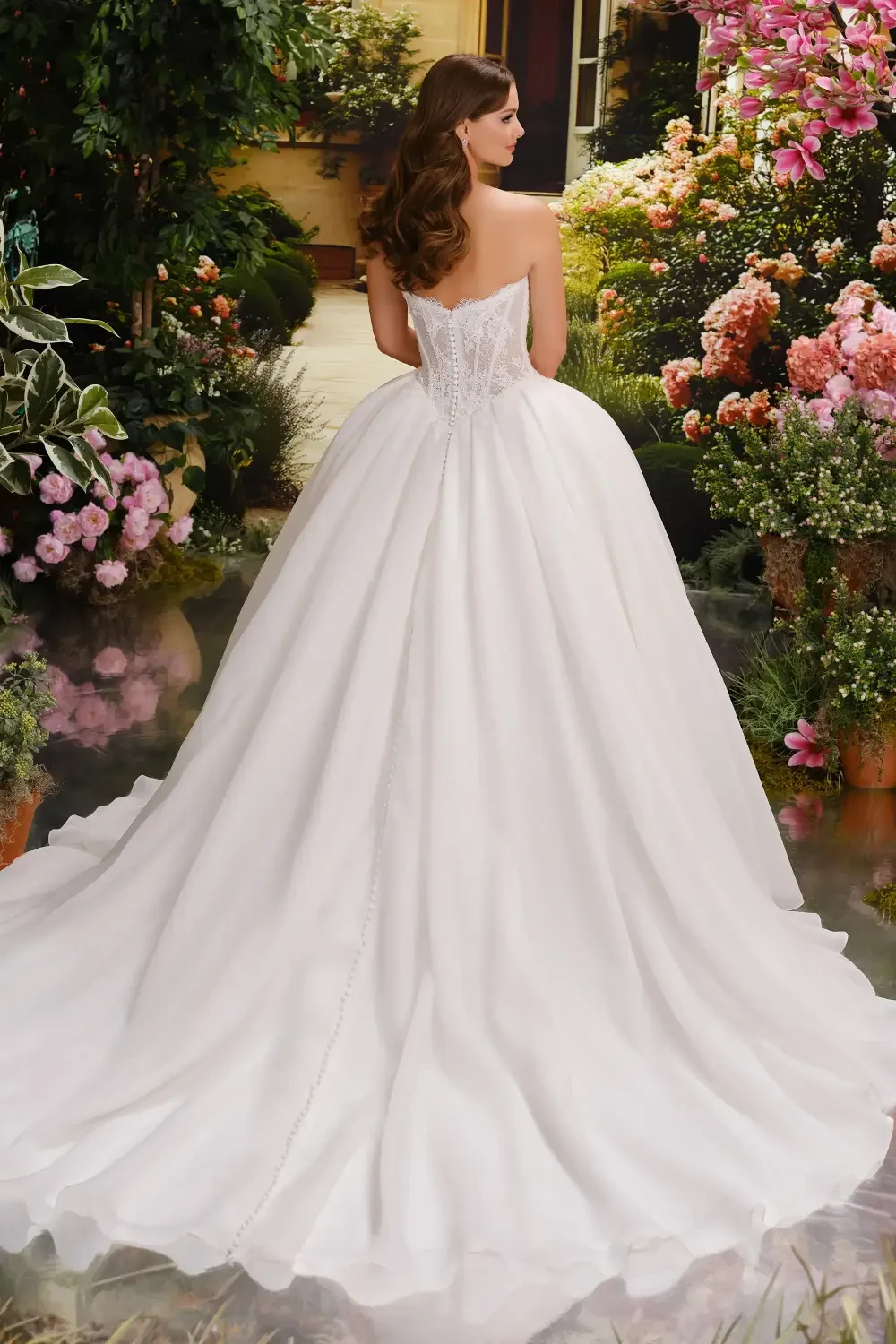 A woman in a strapless white ball gown stands in a garden filled with flowers, smiling confidently.