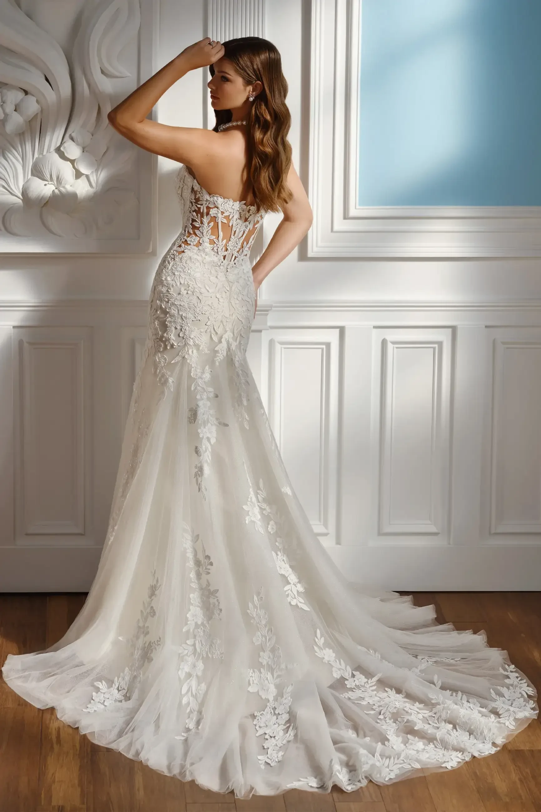 Bridal gown with lace detailing on a woman facing away, showcased in a room with ornate white paneling. Light flows through a large blue window, creating an elegant, serene atmosphere.