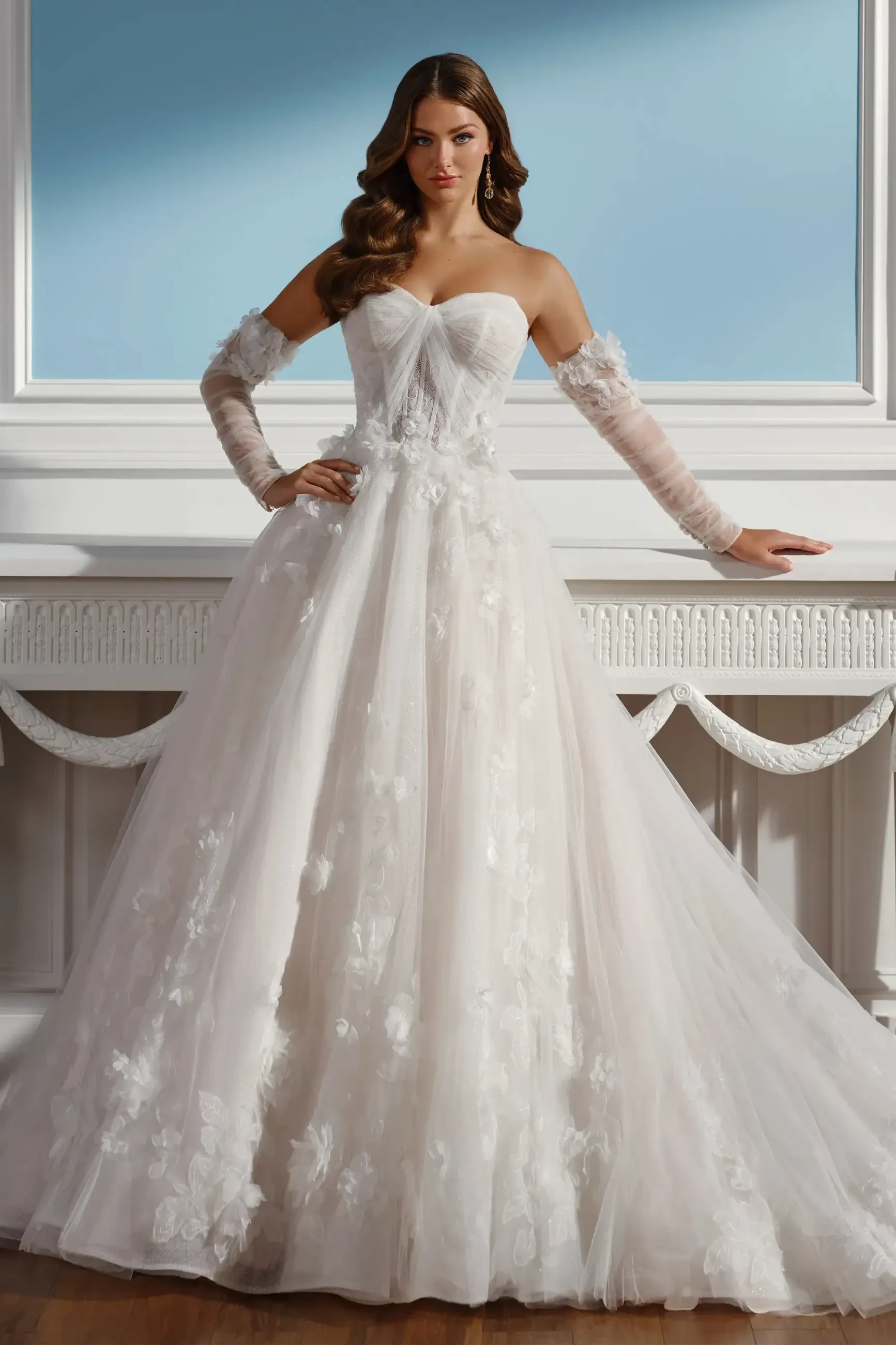 Elegant bride in a strapless, floral lace ball gown, with long sheer gloves, stands confidently by a white ornate mantle against a light blue wall.