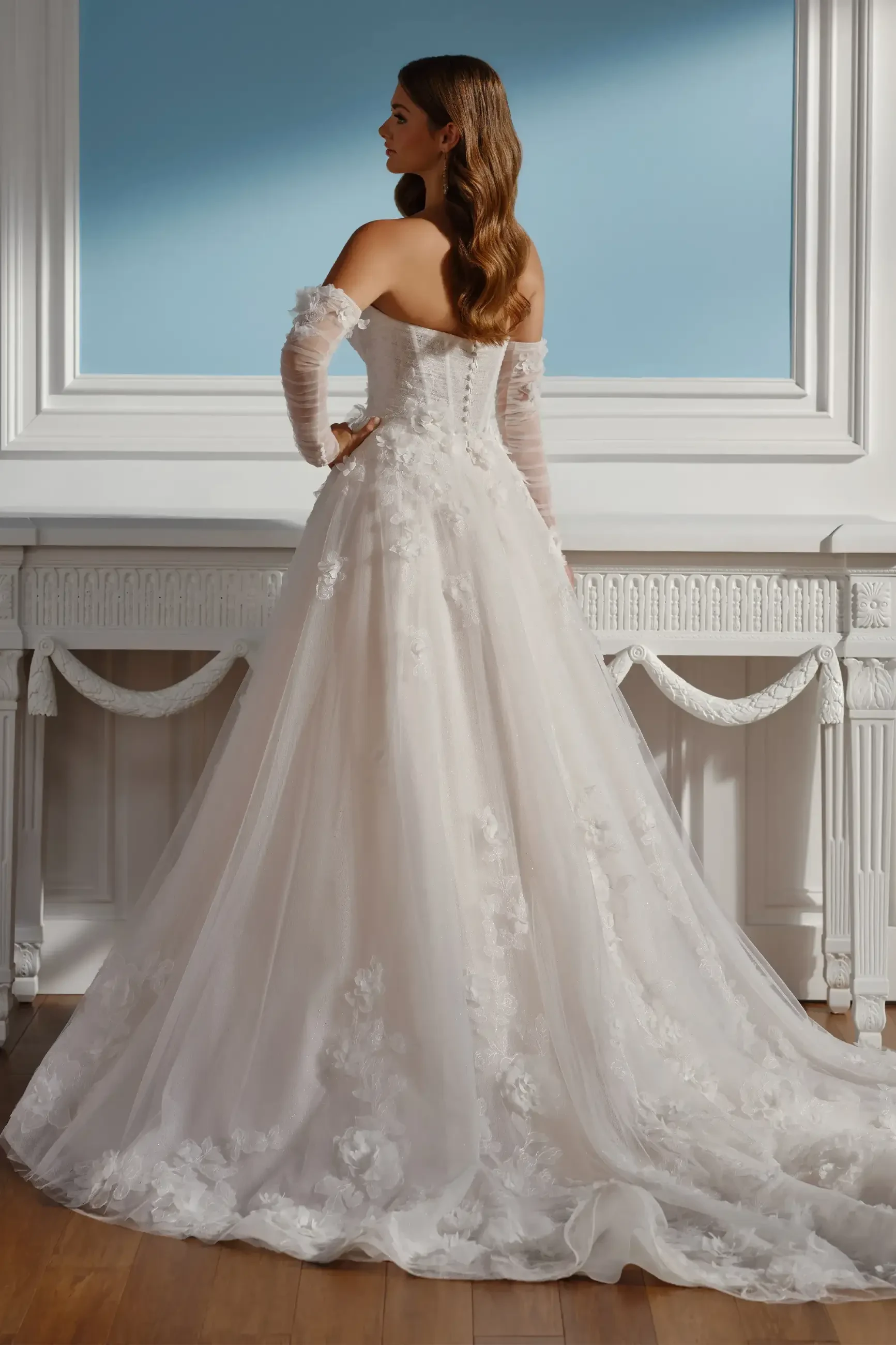 Elegant bride in a strapless, floral lace ball gown, with long sheer gloves, stands confidently by a white ornate mantle against a light blue wall.