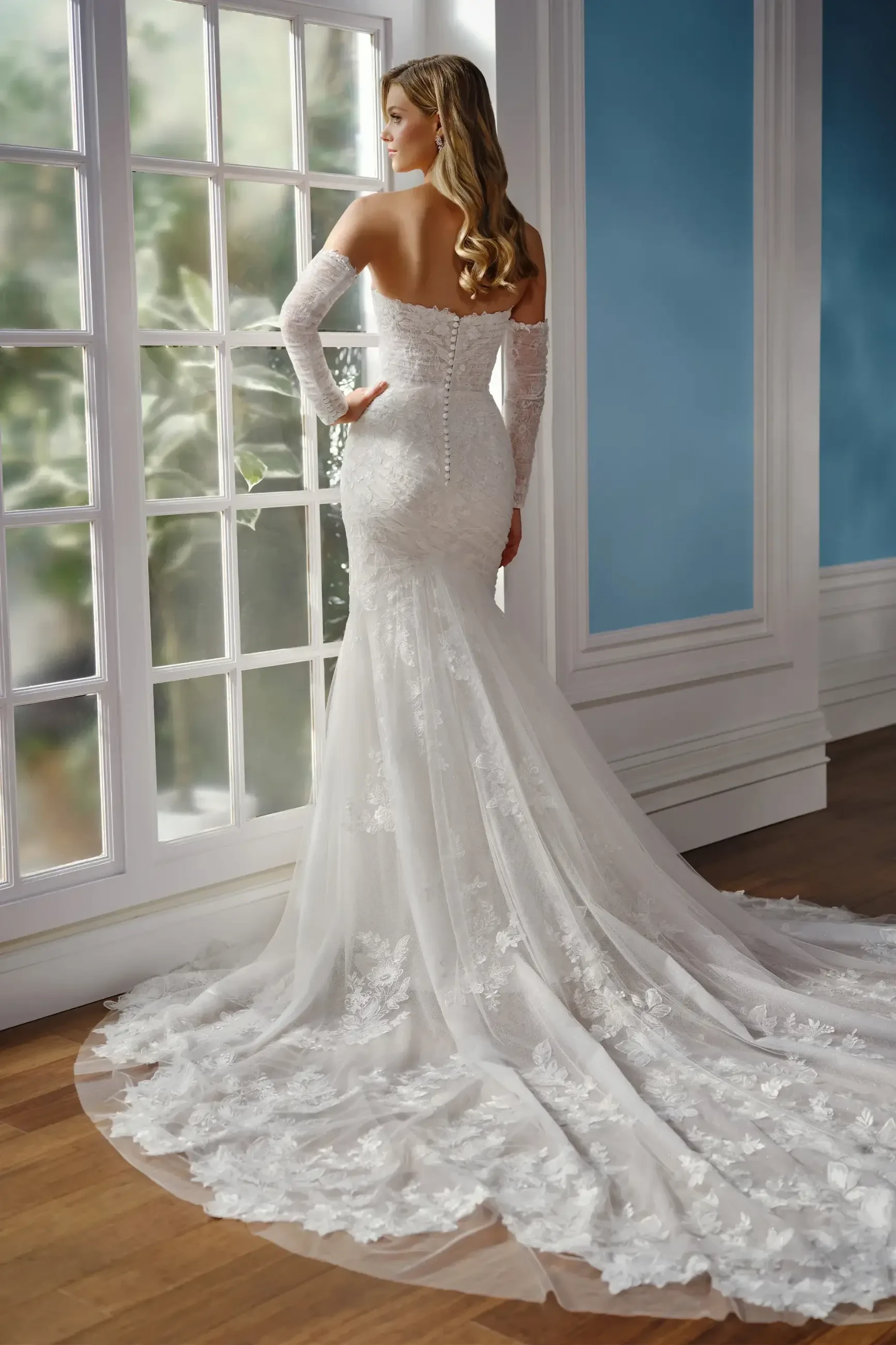 Bride in elegant strapless lace wedding gown with long lace sleeves stands by sunlit window, conveying a serene and romantic atmosphere.