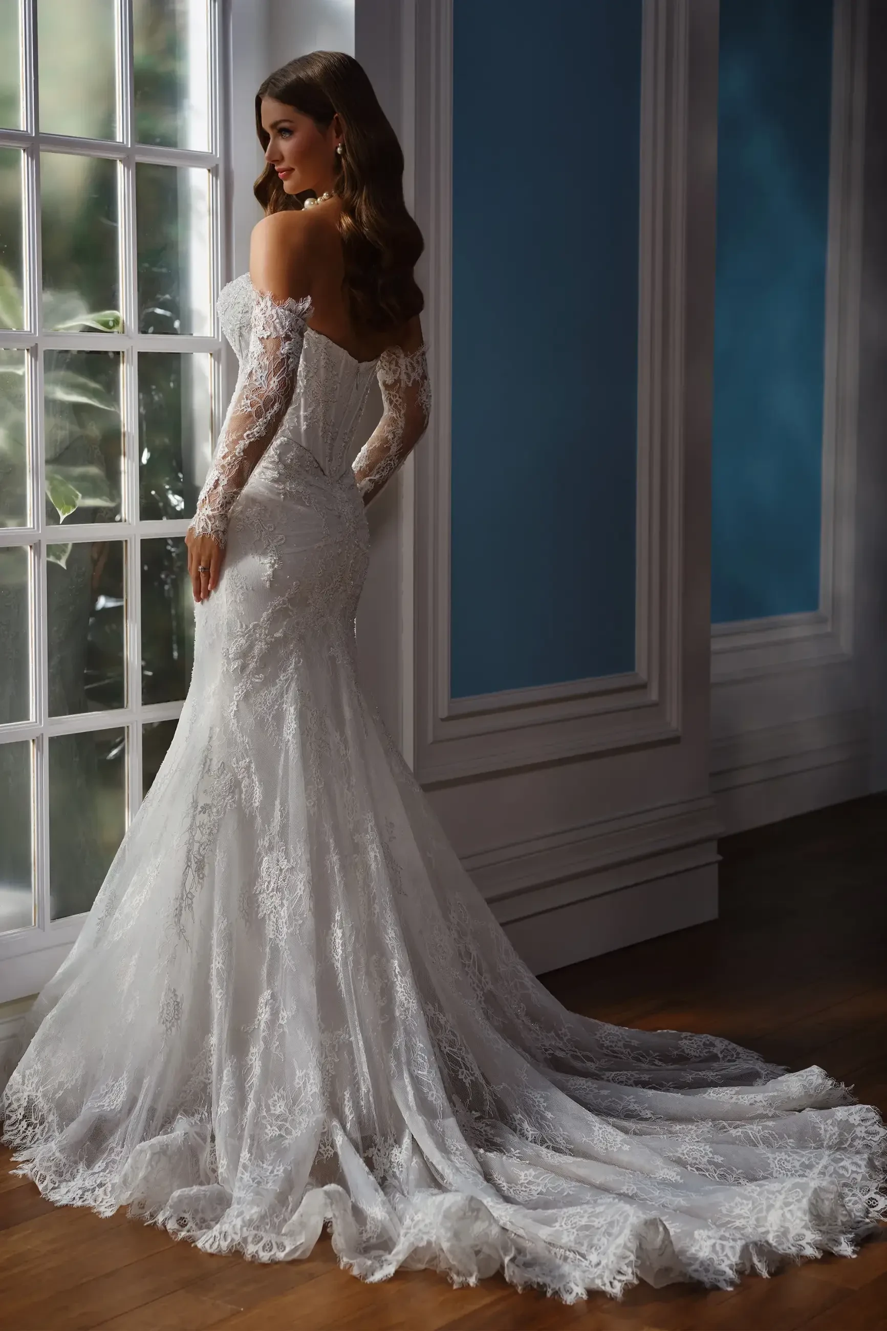 A bride in an off-shoulder lace wedding dress stands by a window. The long train flows elegantly on a wooden floor, creating a romantic and serene mood.