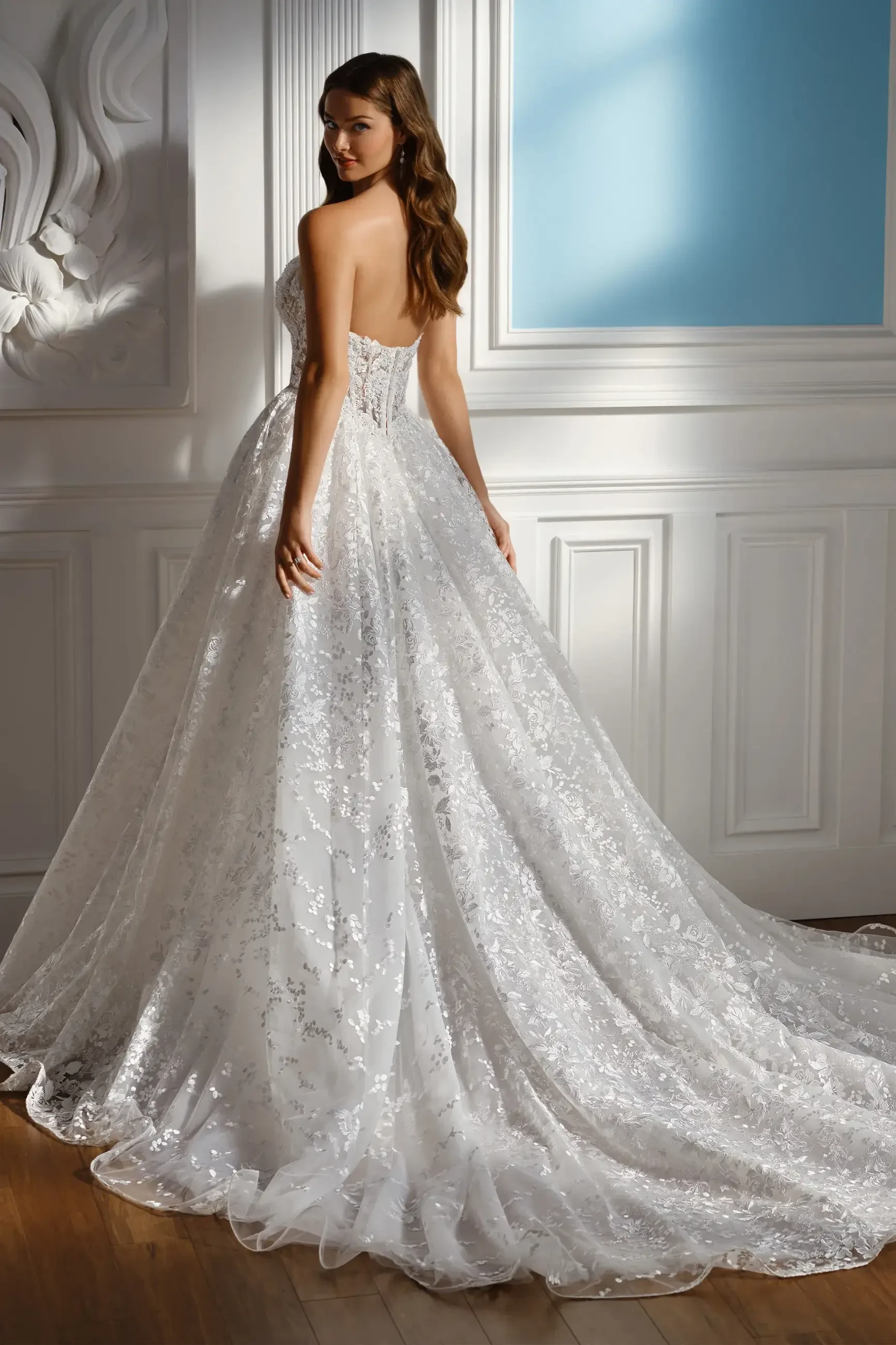 A bride stands gracefully in a backless, lace wedding gown with a flowing train. She poses in a sunlit room, exuding elegance and serenity.