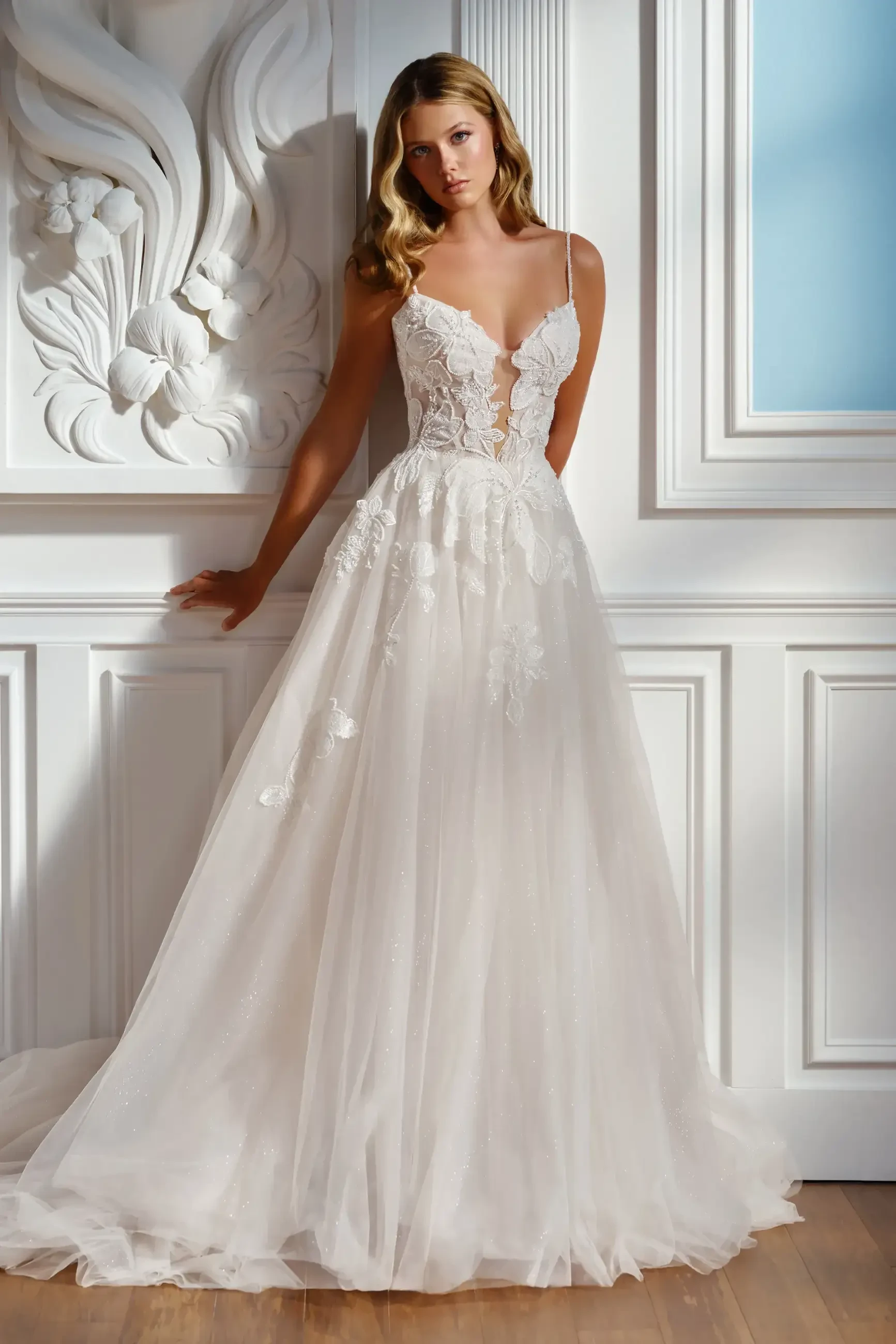 A woman in an elegant, lace wedding dress stands against an ornate white wall. The gown features floral details and a flowing skirt, evoking a romantic ambiance.