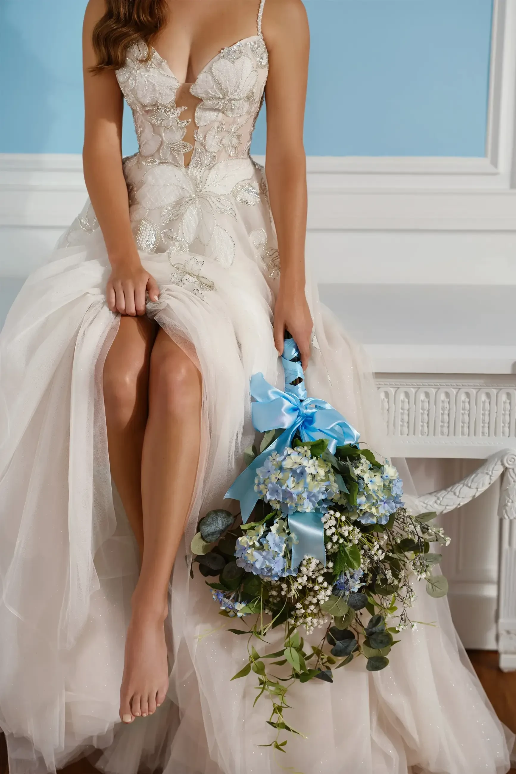 A woman in an elegant, floral lace white gown sits on a white bench, holding a bouquet of blue hydrangeas. The scene is serene with soft blue walls.