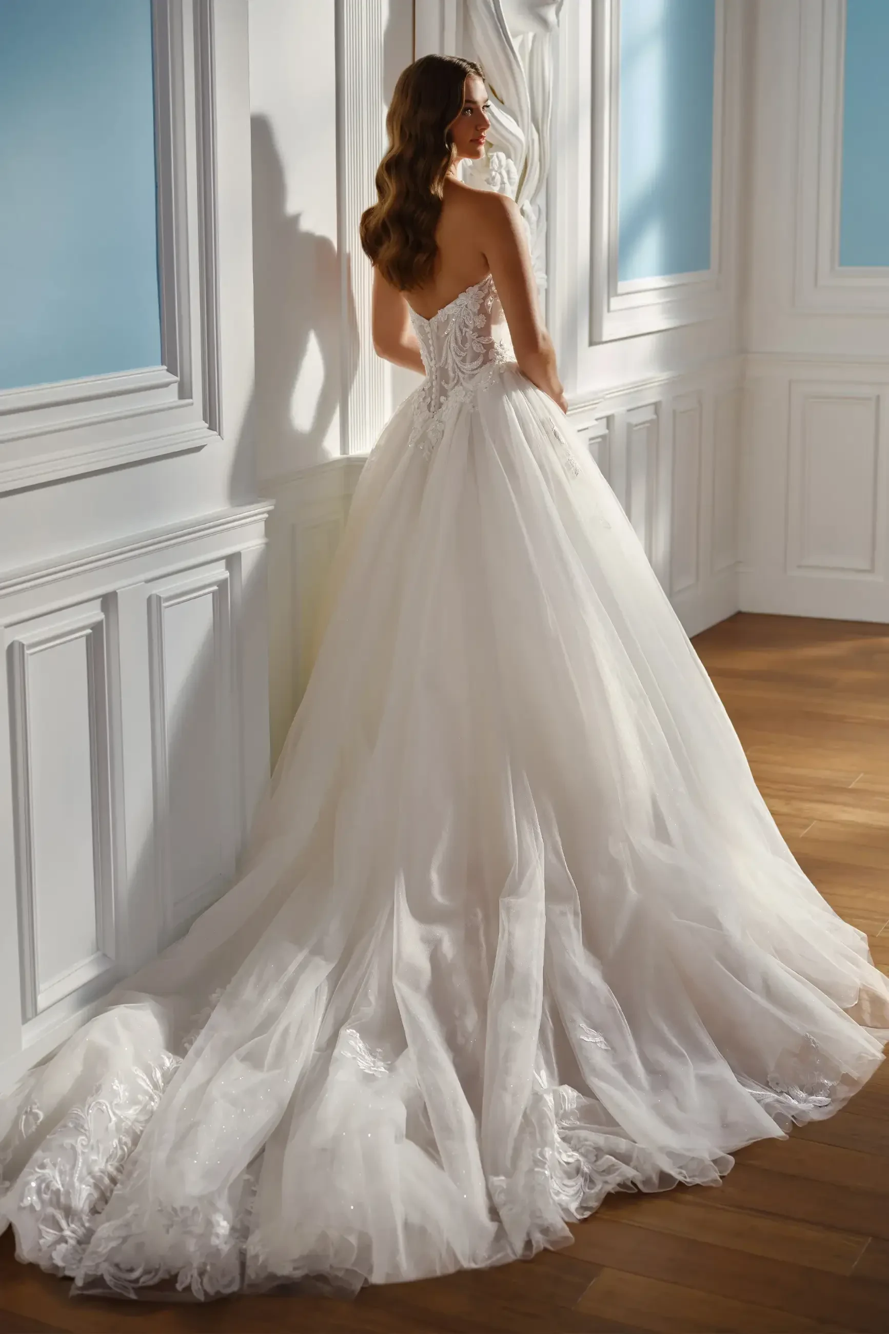 A bride in a strapless white gown stands in an elegant room. The dress has a flowing train, soft light creates a serene mood, and wooden floors add warmth.