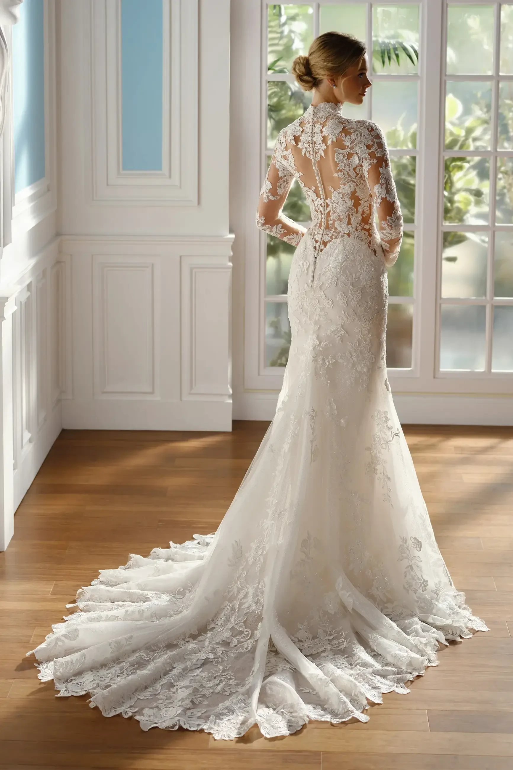 A woman stands by a large window wearing an elegant, backless lace wedding gown with a long train. The room has a serene ambiance, with soft lighting illuminating the wooden floor.