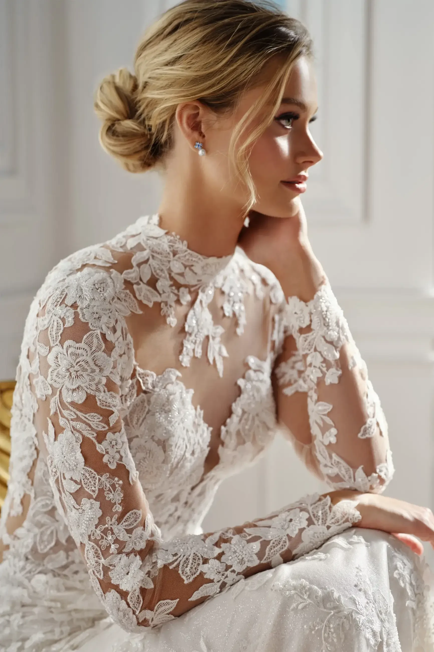 A woman in a lace wedding dress with floral patterns sits gracefully, gazing sideways. Her blonde hair is styled in an elegant bun, exuding a serene, romantic vibe.