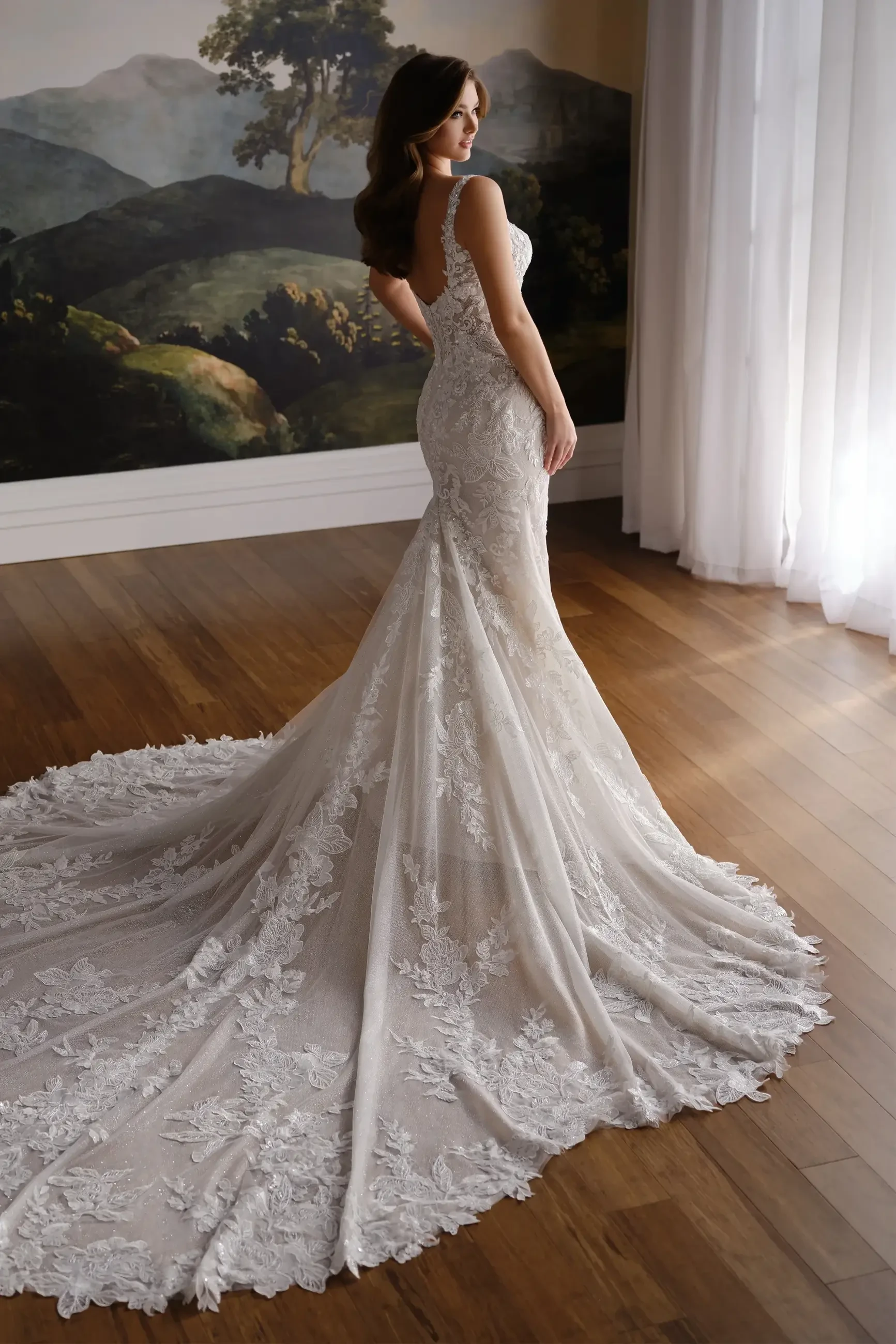 A woman wearing a fitted, lace wedding dress with a long train, standing in front of a large window with natural light. The background features a scenic mural of mountains and trees.
