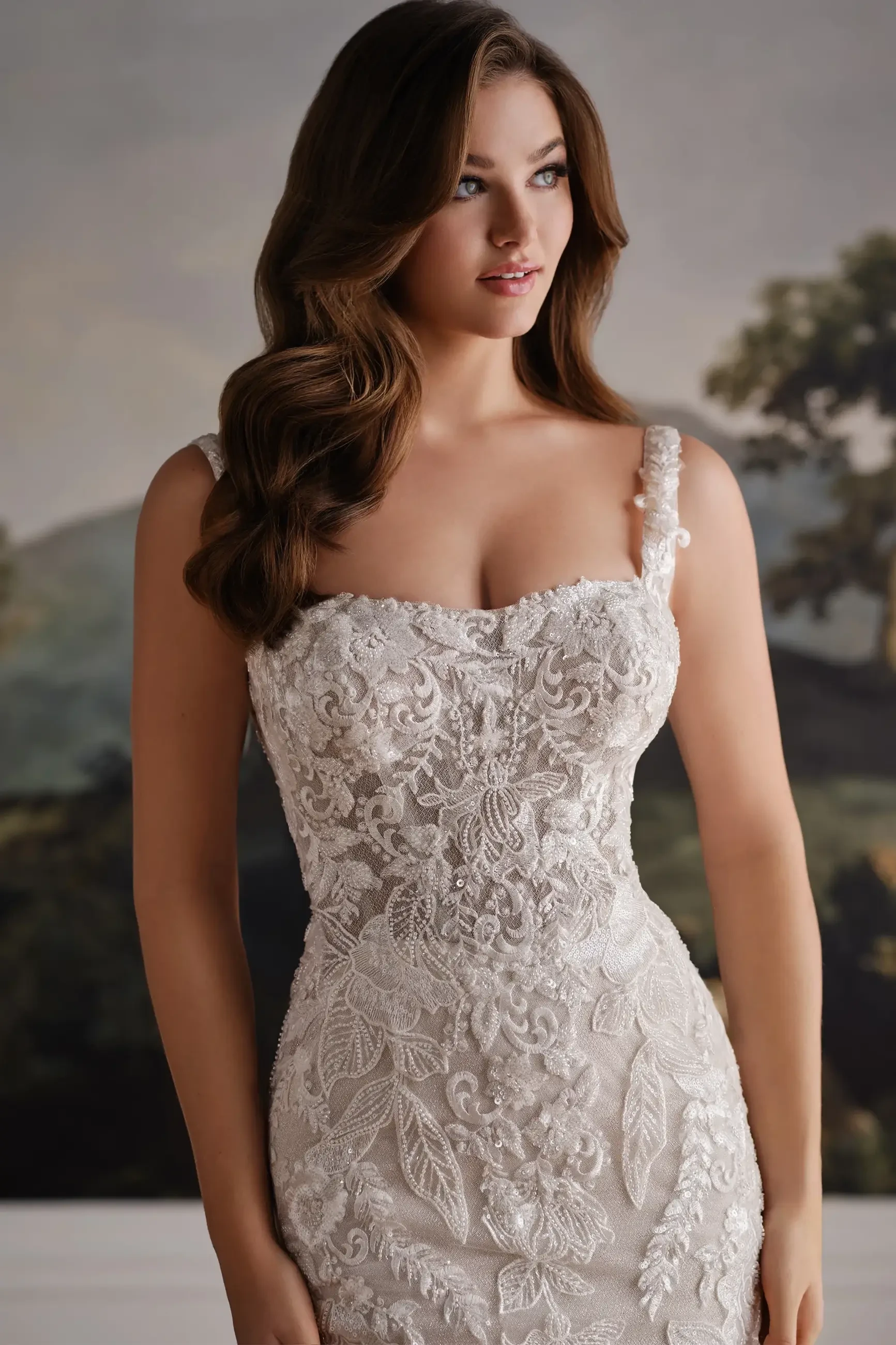 A woman in a detailed lace wedding dress with a sweetheart neckline and floral embroidery, standing against a soft-focus background.