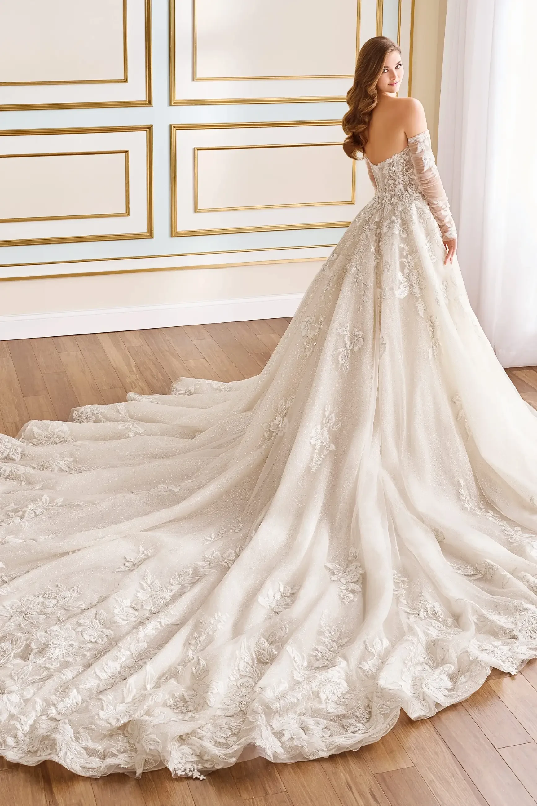 Model is wearing a ball gown with a sweetheart neckline, floral lace appliqués, sequins, glitter net, a dropped waist, Cathedral train, and separate sheer illusion long sleeves.